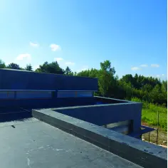 Nowoczesny budynek z płaskim dachem, na którym trwa membrana EPDM montaż. W tle zielony las i błękitne niebo.