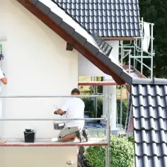 Dwóch mężczyzn na rusztowaniu wykonuje malowanie elewacji domu. Jeden stoi, drugi klęczy, obaj używają wałków.