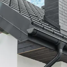 Nowoczesne rynny metalowe kwadratowe w kolorze grafitowym, zamontowane na dachu z ciemną dachówką, obok ceglanego komina.