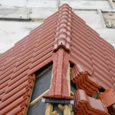 Nowy dach z brązowej dachówki. Widać miejsce przecięcia dwóch połaci dachowych, z elementami wentylacyjnymi i ułożonymi stosami dachówek.