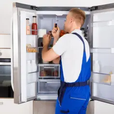 Serwisant w niebieskim kombinezonie sprawdza śrubokrętem wentylator w lodówce.