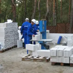 Budowa domu z bloczków betonowych. Pracownicy w niebieskich kombinezonach i białych kaskach przygotowują materiał. Ile bloczków betonowych na m2?