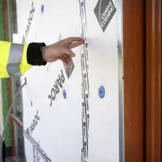 Ręka w żółtej kamizelce wskazuje na taśmę na materiale budowlanym. To kluczowy element wiatroizolacji, chroniący budynek przed wiatrem i wilgocią.