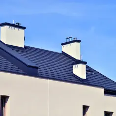 Dach bezokapowy detal: ciemny dach z dachówkami, białe kominy i jasna ściana budynku na tle błękitnego nieba.