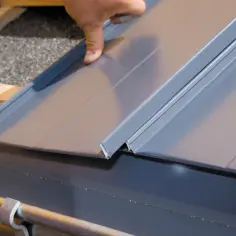 Dłoń montuje szary panel blachy na rąbek na dachu, nad rynną.