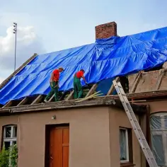 Ekipa na dachu rozkłada niebieską plandekę, przygotowując się do prac. Łacenie dachu cena może być różna, ale bezpieczeństwo jest najważniejsze.