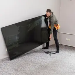 Mężczyzna z pasem narzędziowym przy ścianie trzyma duży, czarny telewizor. Zastanawia się, na jakiej wysokości telewizor będzie najlepiej wyglądał.