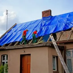 Ekipa na dachu rozkłada niebieską plandekę, przygotowując się do prac. Łacenie dachu cena może być różna, ale bezpieczeństwo jest najważniejsze.