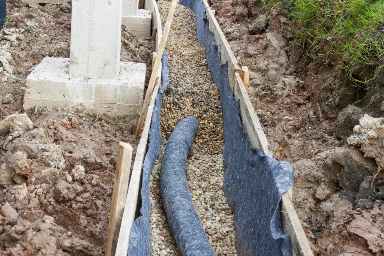 Budowa odwodnienia domu: rura drenarska w wykopie, otoczona żwirem i geowłókniną, przy fundamencie.